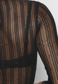 Haut transparent noir à manches longues avec des rayures verticales et un subtil éclat. Présente un motif texturé et une coupe ajustée, révélant des détails de la couche inférieure.