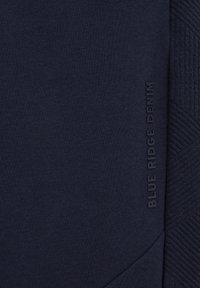 Tumši zils krekls ar teksturētu sānu paneļa un izcelta "BLUE RIDGE DENIM" zīmola logo priekšpusē, lai pievienotu papildu detalizāciju.