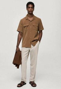 Korte mouwen bruine shirt met twee borstzakken, gecombineerd met lichtbeige broek. Model houdt een bruine gebreide cardigan vast en draagt zwarte sandalen.