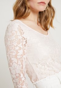 Långärmad, figurnära topp i mjuk rosa spets med blommönster. Har en bred halsringning och genomskinlig textur, perfekt för lager på lager.