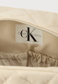 Beigenvärinen tikattu kangas sisäpuolella, jossa kerätyt reunat ja ommeltu etiketti, jossa lukee "Calvin Klein Jeans" mustalla ja sinisellä fontilla. Hopeanvärinen vetoketju.