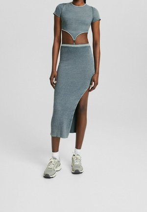 Žena oblečená v modrošedém žebrovaném dvoudílném outfitu s krátkým topem a midi sukní s vysokým rozparkem, doplněném bílými ponožkami a teniskami.