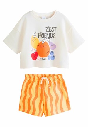 Krótki rękaw jasnokremowej bluzki crop z graficznym motywem owoców i napisem "ZEST FRIENDS FOREVER". Połączona z pomarańczowymi szortami z falistymi paskami.
