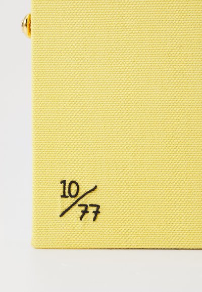 Sac galben din textil, cu o suprafață texturată, având un detaliu brodat în negru "10/77" pe lateral.