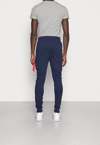 Marineblaue Jogginghosen mit Slim-Fit, mit roten Seitensch Stripes, elastischem Bund und Bündchen. Kombiniert mit einem grauen T-Shirt mit kurzen Ärmeln.