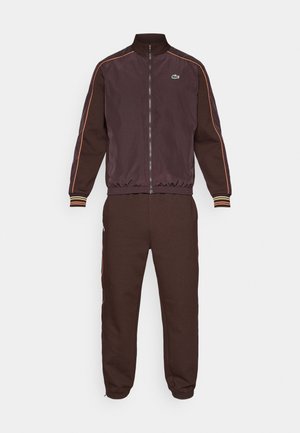 Chándal marrón con chaqueta con cremallera y pantalones a juego. La chaqueta tiene un cuello alto, rayas naranjas y un logo verde; los pantalones tienen puños elásticos.