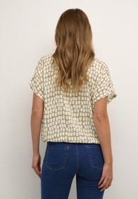 Kortærmet top i kremfarvet med et grønligt geometrisk mønster, parret med blå jeans. Teksturen ser glat ud; designet er afslappet og casual.