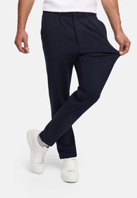 Férfi sötétkék nadrágot és fehér tornacipőt visel, a nadrágszárat nyújtva, hogy bemutassa a szövet rugalmasságát fehér háttér előtt.