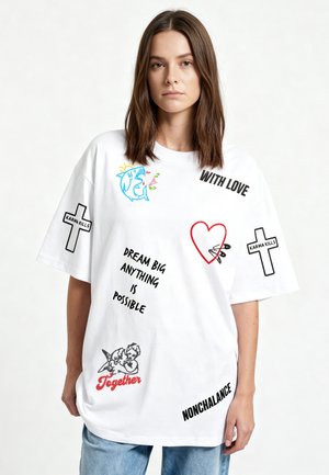 Junge Frau trägt weißes oversized T-Shirt mit bunten Drucken einschließlich Hai, Herz, Kreuzen, Engel und Phrasen wie „Träume groß, alles ist möglich.“