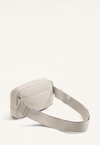 Marsupio in tessuto beige di forma rettangolare, con tasca frontale con cerniera e tracolla regolabile. Texture liscia con dettagli minimali.