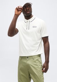 Άνδρας που φοράει λευκό πόλο με μαύρο περίγραμμα και ανοιχτό πράσινο παντελόνι, ρυθμίζοντας λευκό καπέλο-visor ενώ κοιτάζει προς τα πλάγια.