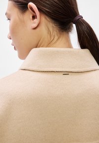 Béžový vlnený kabát s preloženým golierom a jemnou textúrou. Má malú kovovú značku s logom vzadu blízko krčnej línie.