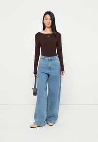 Bodysuit marrón de manga larga con escote cuadrado, combinado con jeans de pierna ancha de color azul claro y un bolso negro. Presenta zapatos planos de color beige.