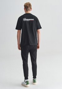 Joven de pie de espaldas con camiseta negra que dice "2022 MODERN" y pantalones negros con un gráfico de estrella en la pantorrilla derecha. Zapatillas blancas.