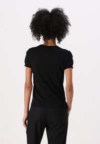 T-shirt nera a maniche corte con scollo rotondo, texture liscia e leggero lucido, abbinata a pantaloni neri. Vista da dietro.
