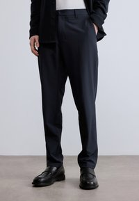 Na mieru šité námornícke nohavice z hladkej látky, so skladaným predným dielom, bočnými vreckami a klasickým strihom, doplnené čiernymi mokasínmi.