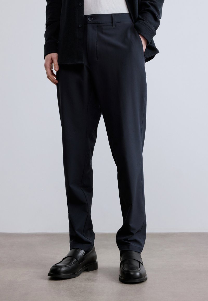 Na mieru šité námornícke nohavice z hladkej látky, so skladaným predným dielom, bočnými vreckami a klasickým strihom, doplnené čiernymi mokasínmi.