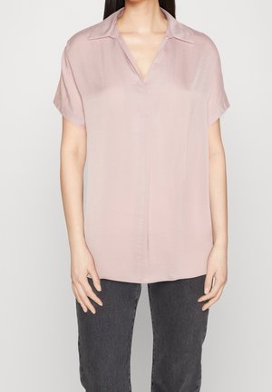 Vrouw draagt een lichtroze loszittende blouse met korte mouwen, een kleine kraag en een V-hals, gecombineerd met donkergrijze jeans.