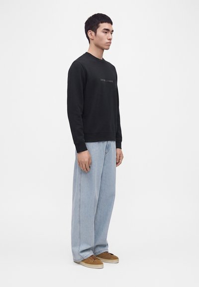 Jeune homme debout, portant un sweat-shirt noir, un jean large bleu clair et des baskets marron, face à droite sur un fond blanc uni.