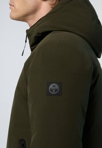 Olivenfarvet pufferjakke med hætte, glat tekstur og lappe med logo på venstre ærme. Lynlåslommer er synlige på siden.