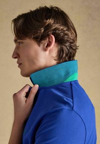 Jeune homme ajustant le col relevé d'un polo bleu avec une doublure intérieure rayée verte et bleue, vu de profil.