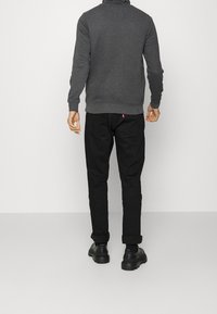 Sweat-shirt gris à col montant, jeans noirs et chaussures noires. Le sweat-shirt est doté de poignets et d'un ourlet côtelés. Branding subtil sur les jeans.