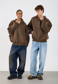 Rjavi zip-up kapuci s oversized dizajnom; model s lijeve strane ima pletenice. Labave traperice—tamno plave s lijeve strane, svijetlo plave s desne strane, uz crne cipele.