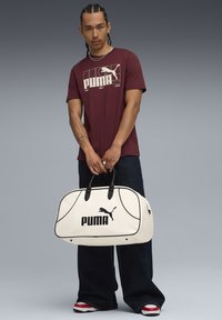 Mørkerød t-shirt med hvid PUMA-logo, mørkeblå brede jeans og en hvid duffelbag med sorte detaljer og logo. Røde og hvide sneakers.