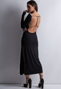 Robe noire à manches longues avec le dos nu, dotée de fines bretelles et d'une fente sur le côté. Texture lisse ; associée à des chaussures à talons hauts noires.