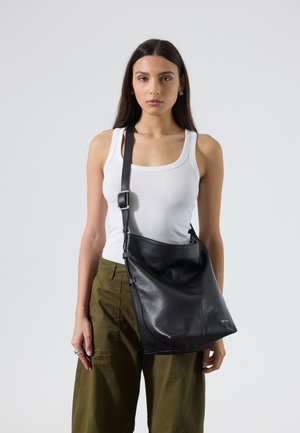 Mujer con pelo largo y oscuro, vestida con camiseta sin mangas blanca y pantalones color oliva, que lleva un gran bolso negro de cuero para hombro con correa ajustable.