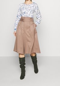 Donna che indossa una gonna beige a vita alta con cintura, una blusa bianca a maniche lunghe con fantasie floreali blu e stivali neri sopra il ginocchio.