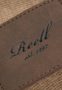 Parche de cuero marrón con bordes cosidos, grabado con el texto en cursiva "Reedl est. 1997" sobre un fondo de tela texturizada.