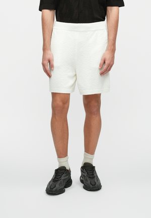Witte textuurshorts met een elastische tailleband, gedragen met zwarte sportschoenen en lichtgrijze sokken; staande tegen een effen achtergrond.
