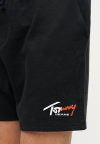 Μαύρα σορτς με λευκό και κόκκινο λογότυπο "Tommy Jeans" στο κάτω δεξί πόδι, φορεμένα από ένα άτομο που στέκεται μπροστά σε μονόχρωμο φόντο.