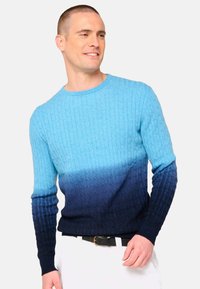 Pull en maille torsadée dégradé bleu et marine avec un col rond. Présente un tissu texturé et des poignets côtelés. Associé à un pantalon blanc.