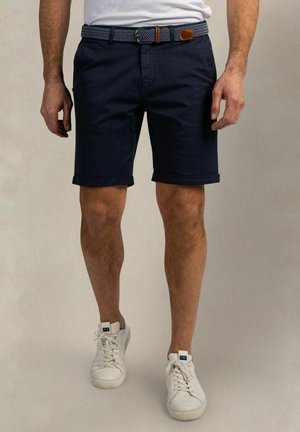 Man draagt donkerblauwe korte broek met een geweven riem, witte sneakers en een wit overhemd ingestopt, staand tegen een effen achtergrond.