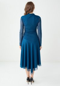 Robe teal avec de longues manches transparentes, bodice ajusté et jupe évasée qui tombe jusqu'aux mi-mollets. Portée avec des chaussures à talons hauts noires.