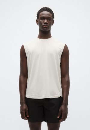 Gaiši bēšs bezroku sporta krekls ar apaļu kakla izgriezumu, izgatavots no gluda, viegla materiāla. Valdāts kopā ar melnām šortām.