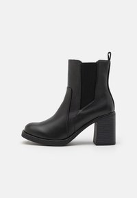 Musta nahkasaapas nilkkapituisena, jossa on tukevat korot, joustavat sivupaneelit ja vetoketju, esiteltynä ulkopuolelta valkoista taustaa vasten.