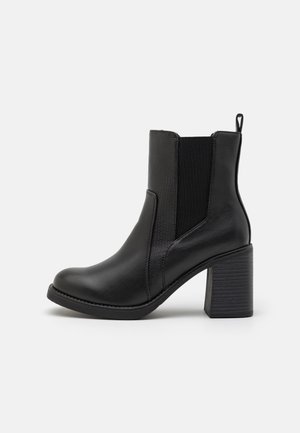 Schwarze Leder-Stiefelette mit Blockabsatz, elastischen seitlichen Einsätzen und Zuglasche, von der Außenseite auf einem weißen Hintergrund gezeigt.