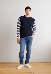 Navy sweater vest over a striped long-sleeve shirt, paired with blue jeans and white sneakers. Wooden floor backdrop, minimalistic design.