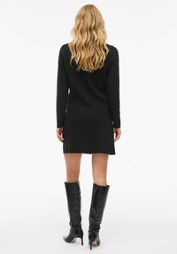 Ensemble en tricot noir comprenant un pull et une jupe ; design ajusté avec des manches longues et un ourlet évasé. Porté avec des bottes en cuir noir montant jusqu'au genou.