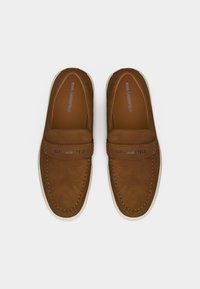 Καφέ μοκασίνια Karl Lagerfeld από σουέντ με λευκές σόλες, ραφές πάνω και μεταλλική λεπτομέρεια με λογότυπο στον ιμάντα, φαίνονται από πάνω.