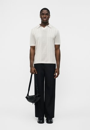 Homme portant un polo en maille texturée beige à manches courtes, un pantalon noir à jambes larges, des chaussures noires, tenant un sac à bandoulière en cuir noir.