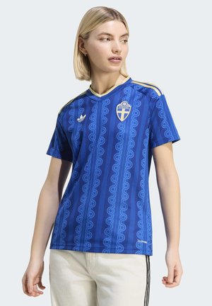 Kvinde iført en blå svensk fodboldtrøje med guldstriber og emblem, kombineret med lyse bukser, der kigger sidelæns.