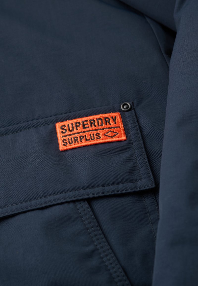 Marineblauwe stof met een rechthoekig oranje stuk genaaid, waarop de tekst "SUPERDRY SURPLUS" en een klein diamantsymbool te zien zijn.