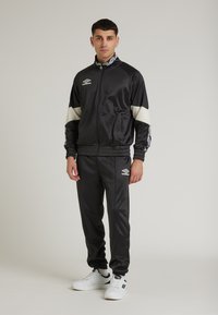 Sort atletisk træningsdragt lavet af skinnende stof; har hvide detaljer på ærmer og ben, jakke med lynlås, elastiske manchetter og logo-detalje.
