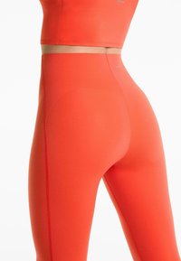 Close-up van een persoon die felle oranje high-waisted legging en een bijpassende crop top draagt, met zichtbare naden en het logo op beide kledingstukken.