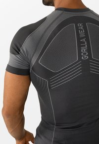 Močan moški, oblečen v tesno črno-sivo majico Gorilla Wear z teksturiranimi vzorci in logotipom na zgornjem delu hrbta, na enobarvnem ozadju.