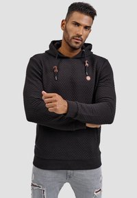 Zwarte gebreide hoodie met een trekkoordkap, voorzien van een klein logo en geribde manchetten. G gedragen met lichtgrijze distressed jeans.